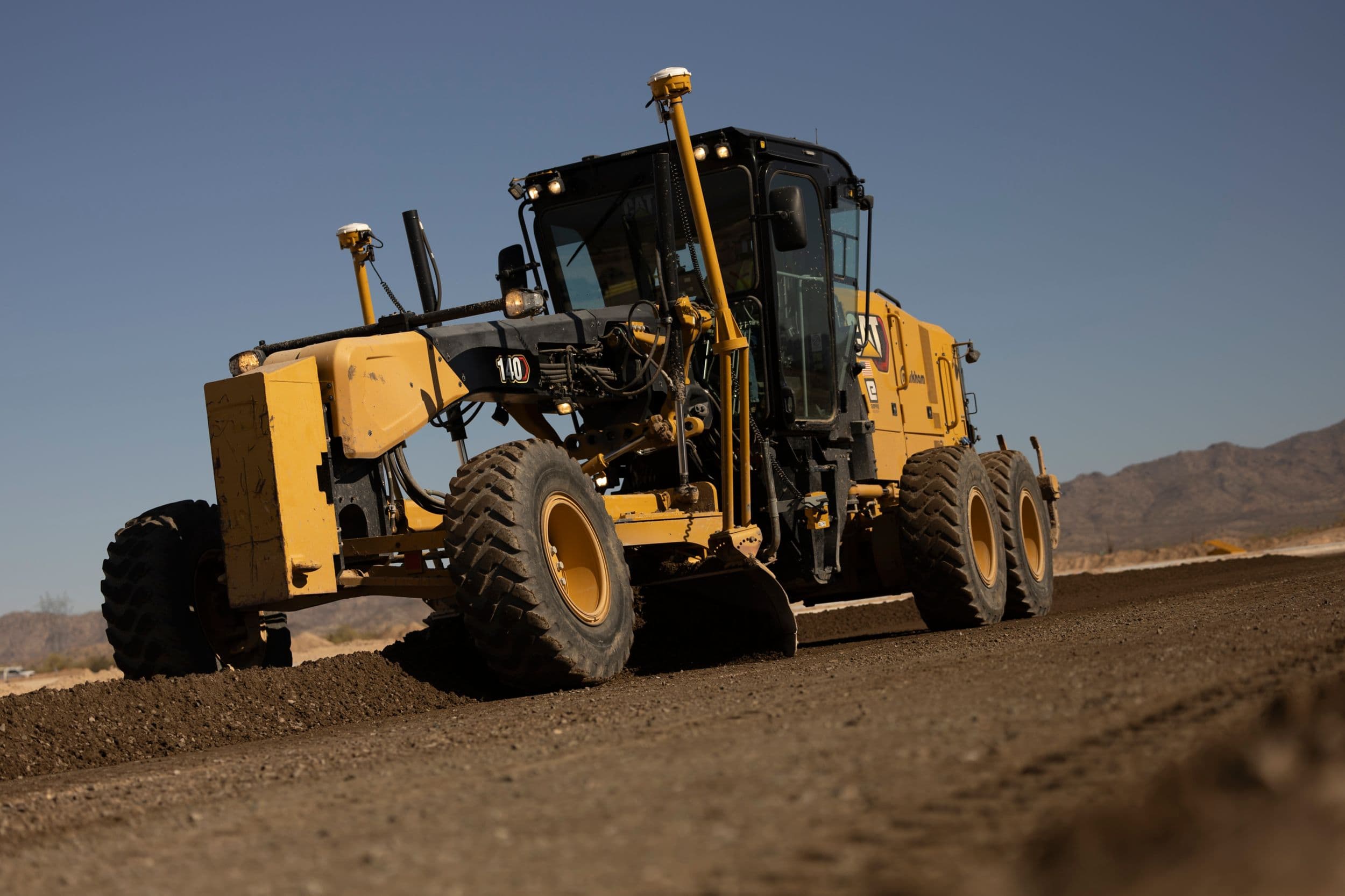 140 Motor Grader at work grading a dark dirt road with concrete curbs installed. This machine has a lever and steering wheel control system.