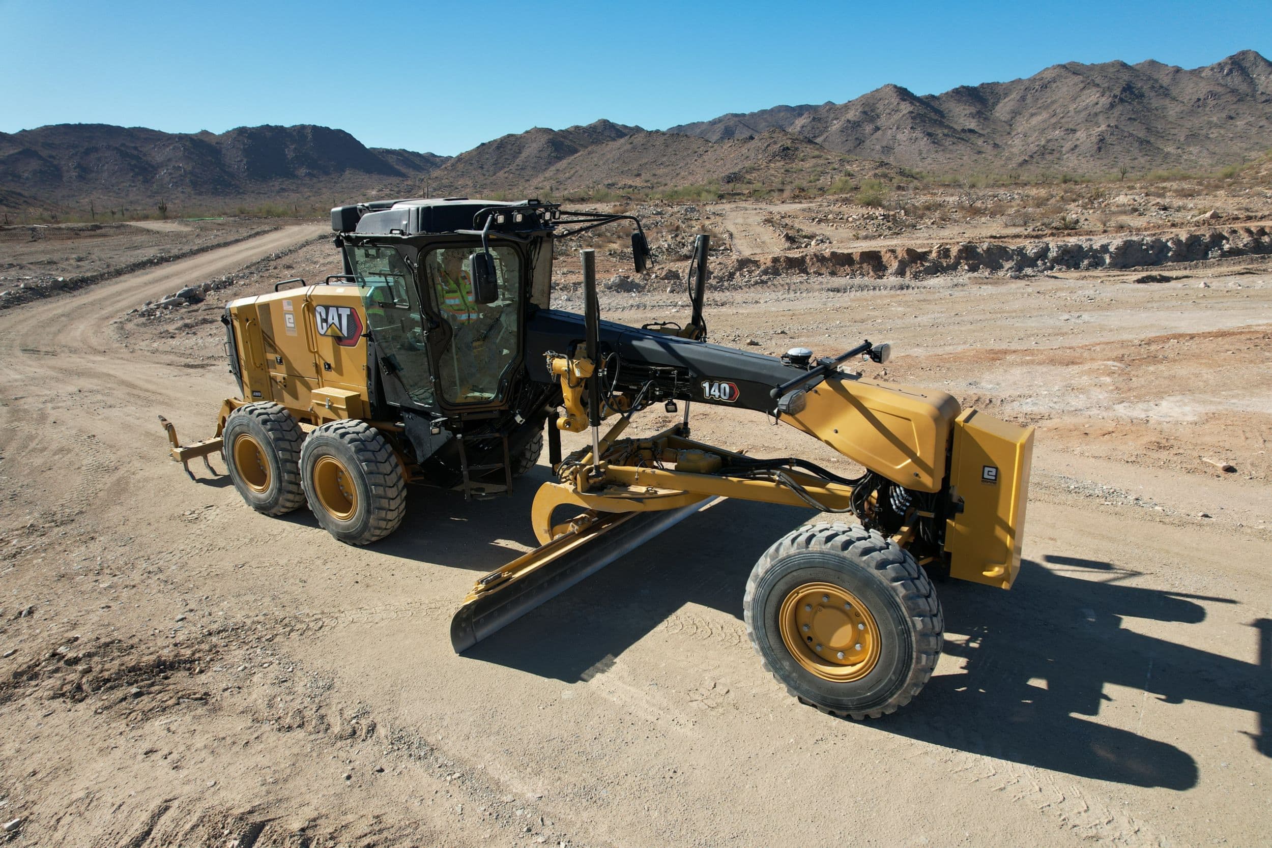 140 Motor Grader in the desert. The machine has the center moldboard resting on the ground and the machine is not in motion. There is a rear ripper, a front counterweight, and a Joystick control configuration.